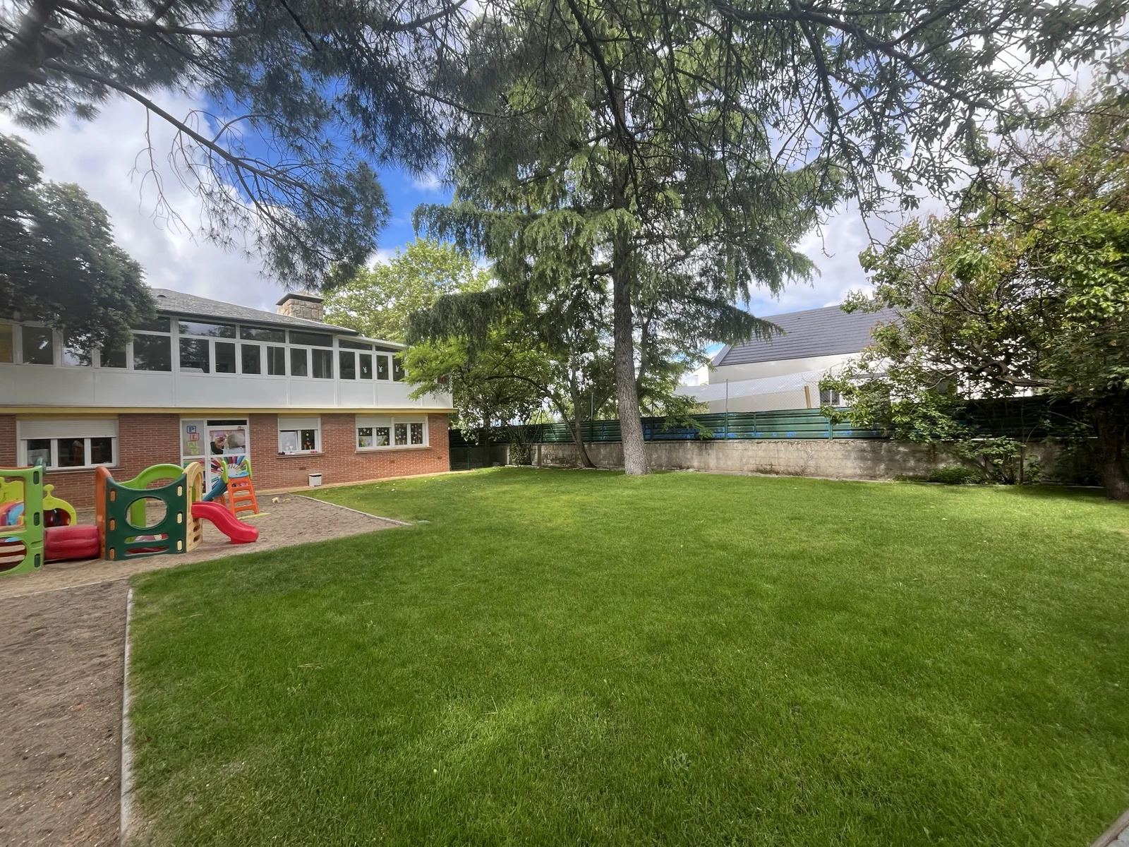 Jardín de Escuela Infantil Cascarón Galapagar, con césped, árboles, zona de juegos y el edificio de la escuela.